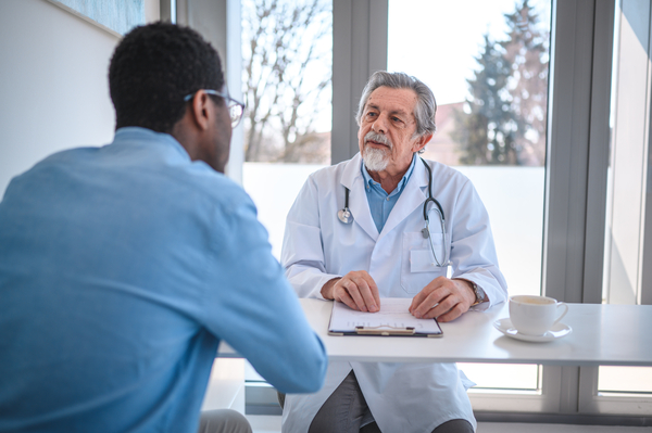 Older provider talking to a middle aged patient at his desk. There is a clipboard and a cup of coffee sitting on desk