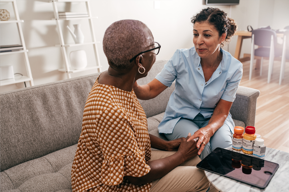 Provider and patient sitting on a couch with pill bottles and a tablet on a table. The meeting is inside the patient's home